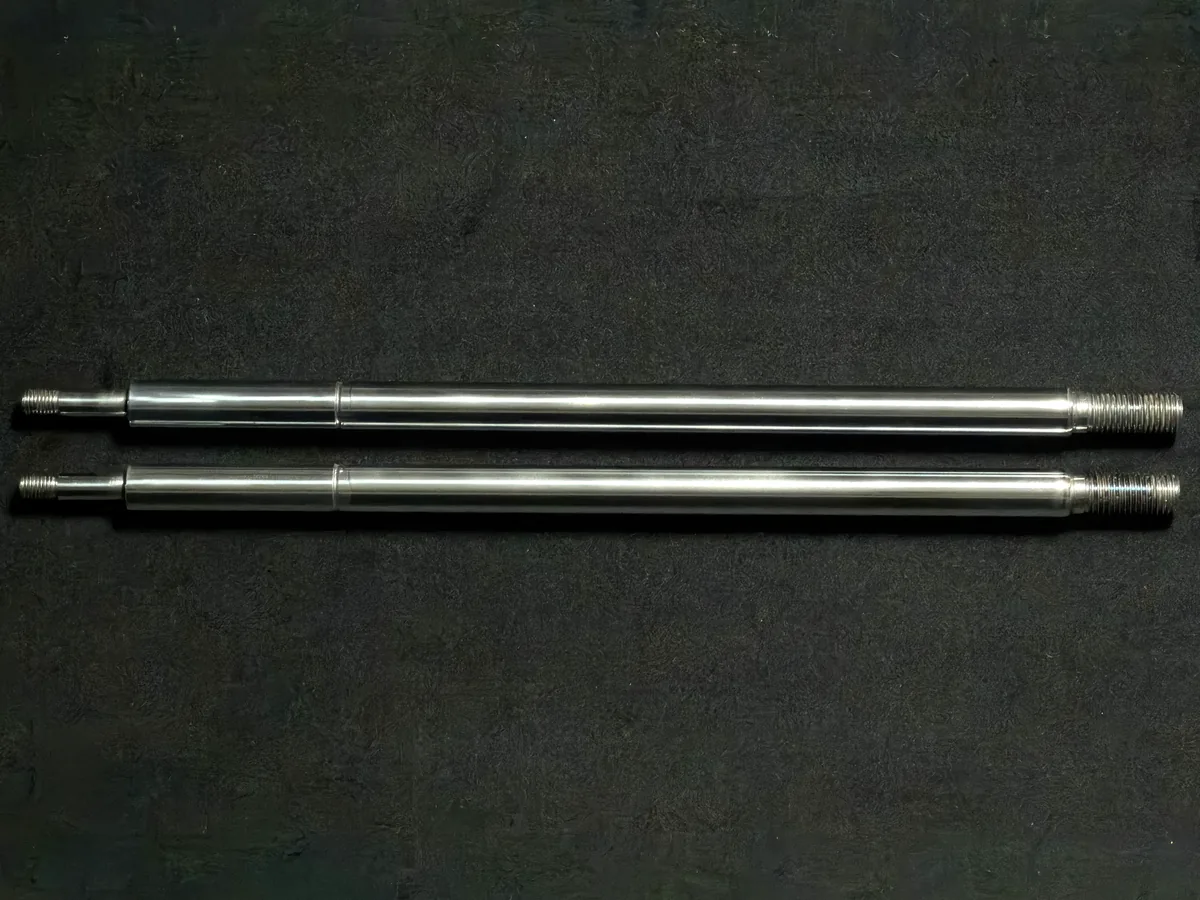 Two TripleHard® coated shock absorber rods lying side by side on a dark surface, threaded ends to the right