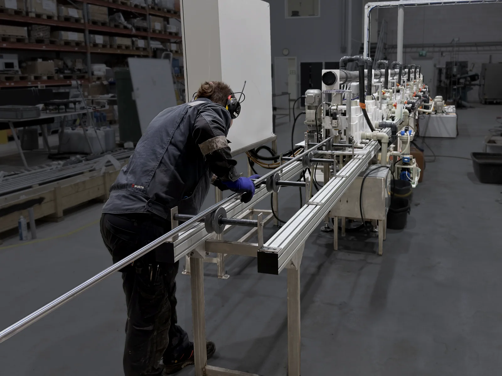 An operator in ear defenders and gloves feeds a hydraulic cylinder rod onto the entry rail of the enclosed TripleHard® line in Kuopio