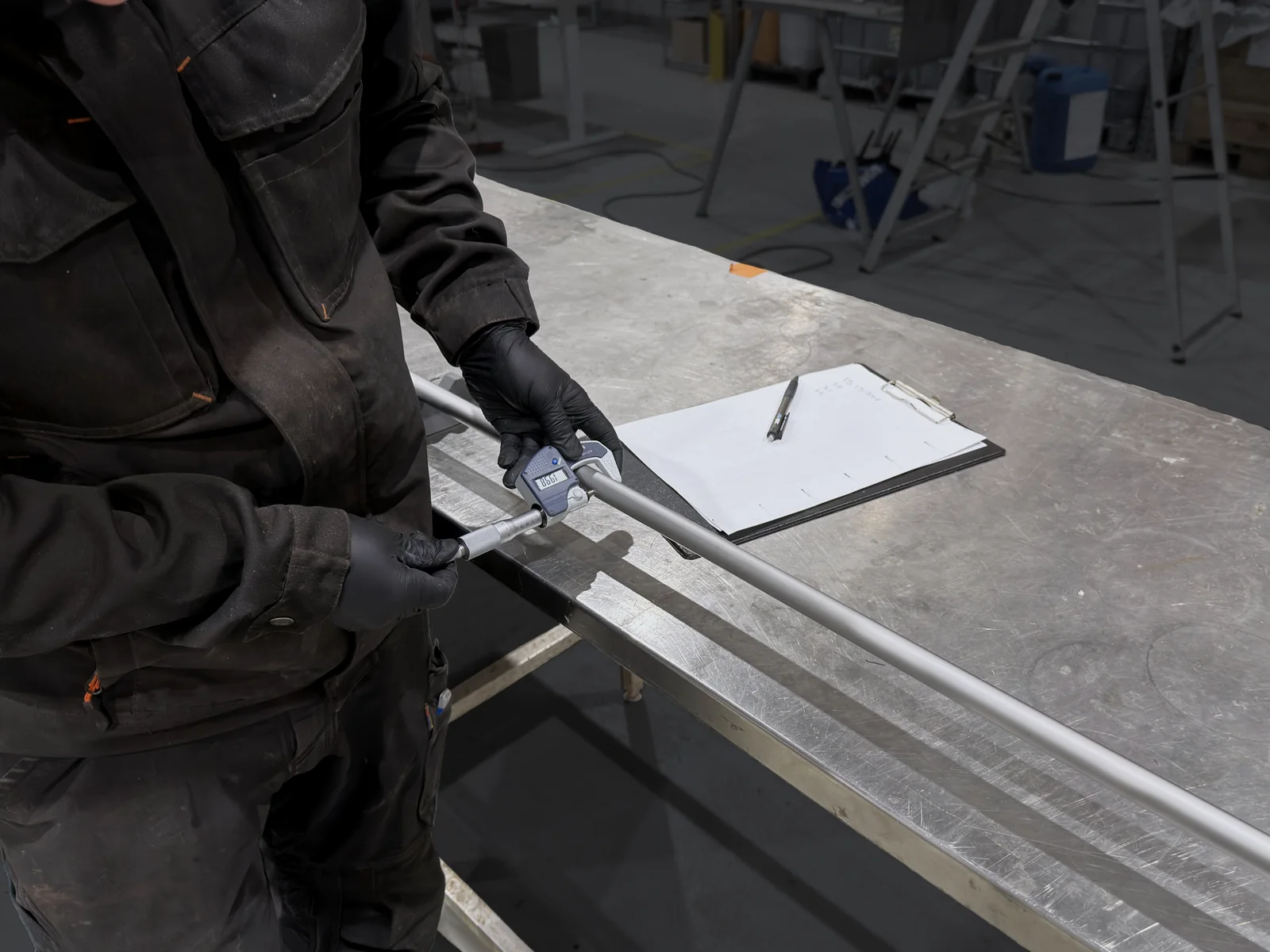 Gloved hands measure the coating on a TripleHard® coated rod with a micrometer at the measuring table, a record sheet beside the rod